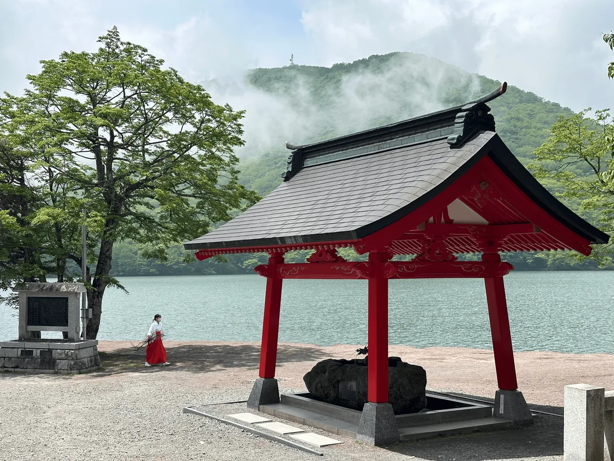 A red temple structure in the foreground, with a maiko shrine maiden dressed in white and red walking along the lake shoreline.