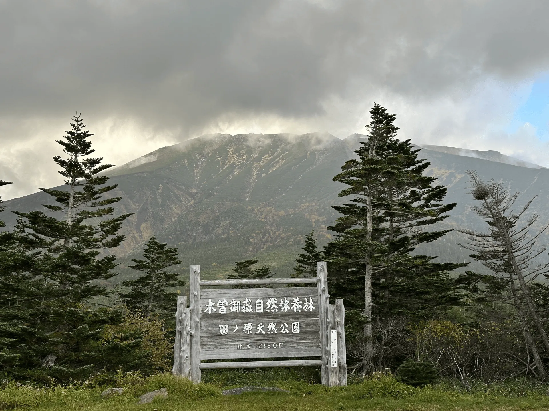 The peak of Mt Ontake, mostly visible. In the front is a wooden sign, marking the area as a nature park.