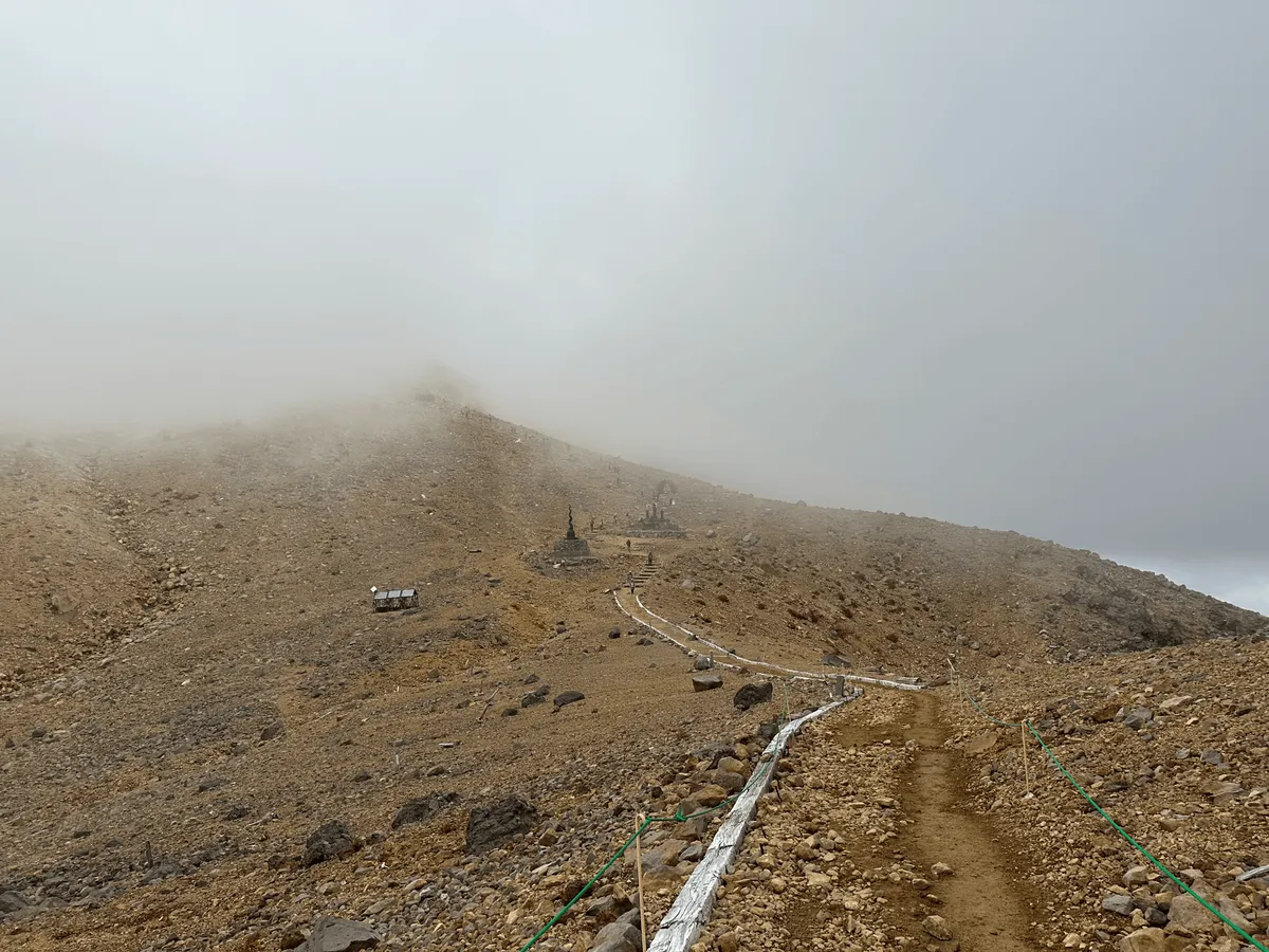 A foggy path up a mountain. It's just covered in tiny beige rocks. There are some small black statues in the distance.