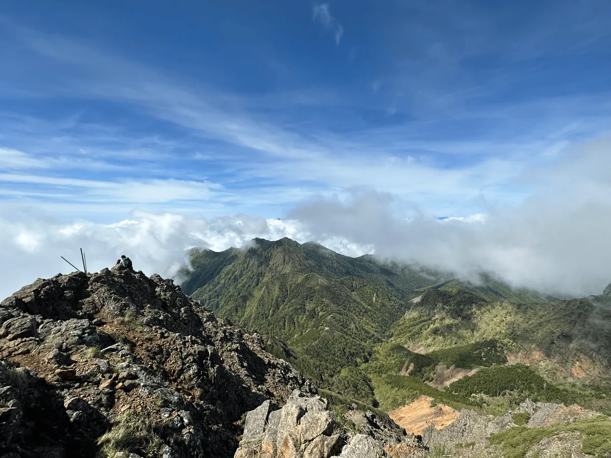 In the foreground, the rocky summit of Mt Aka. In the background, blue skies, fog and green mountains.